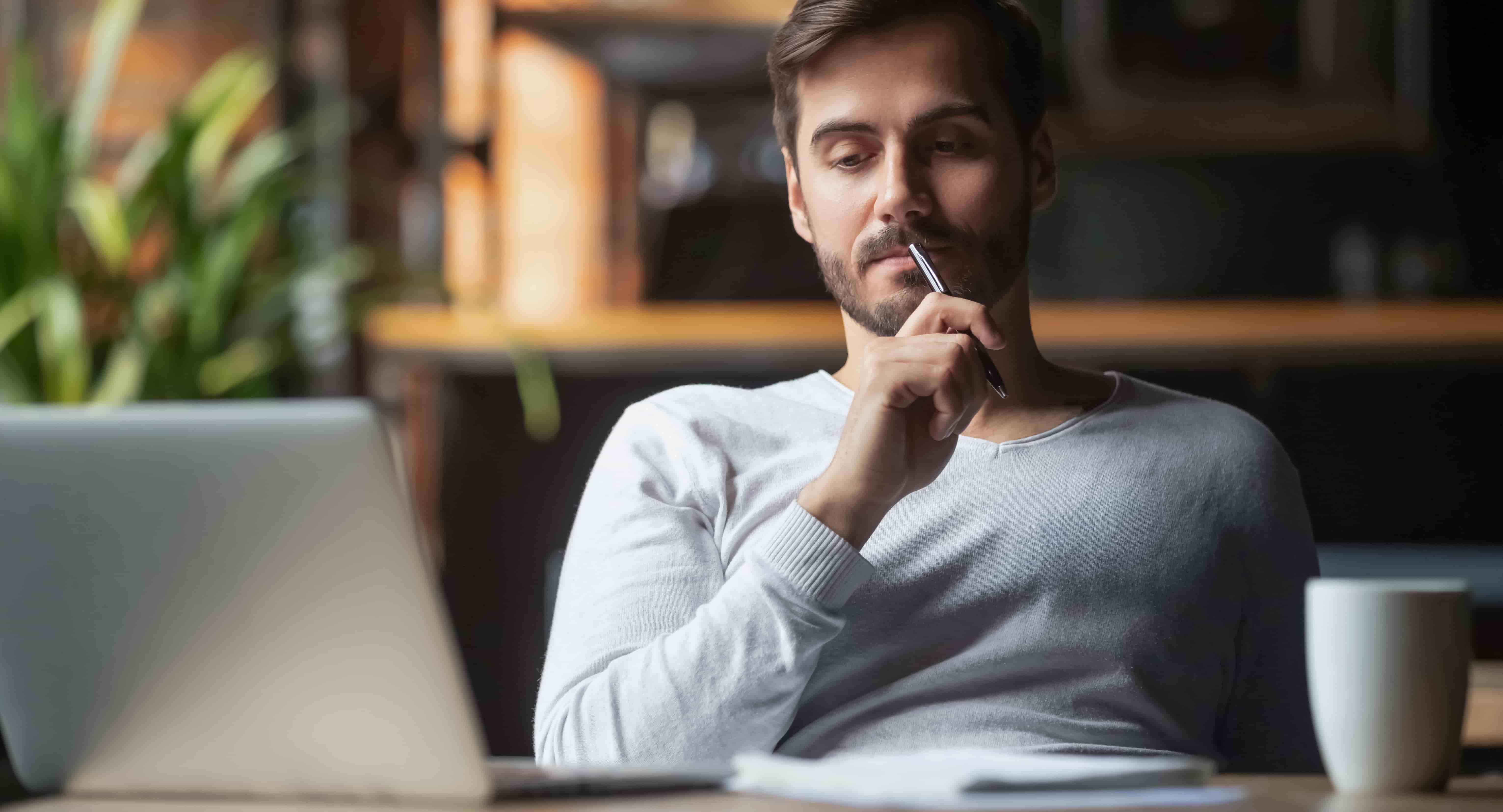 Pensive bearded man sitting at table drink coffee work at laptop thinking of problem solution, thoughtful male employee pondering considering idea looking at computer screen making decision